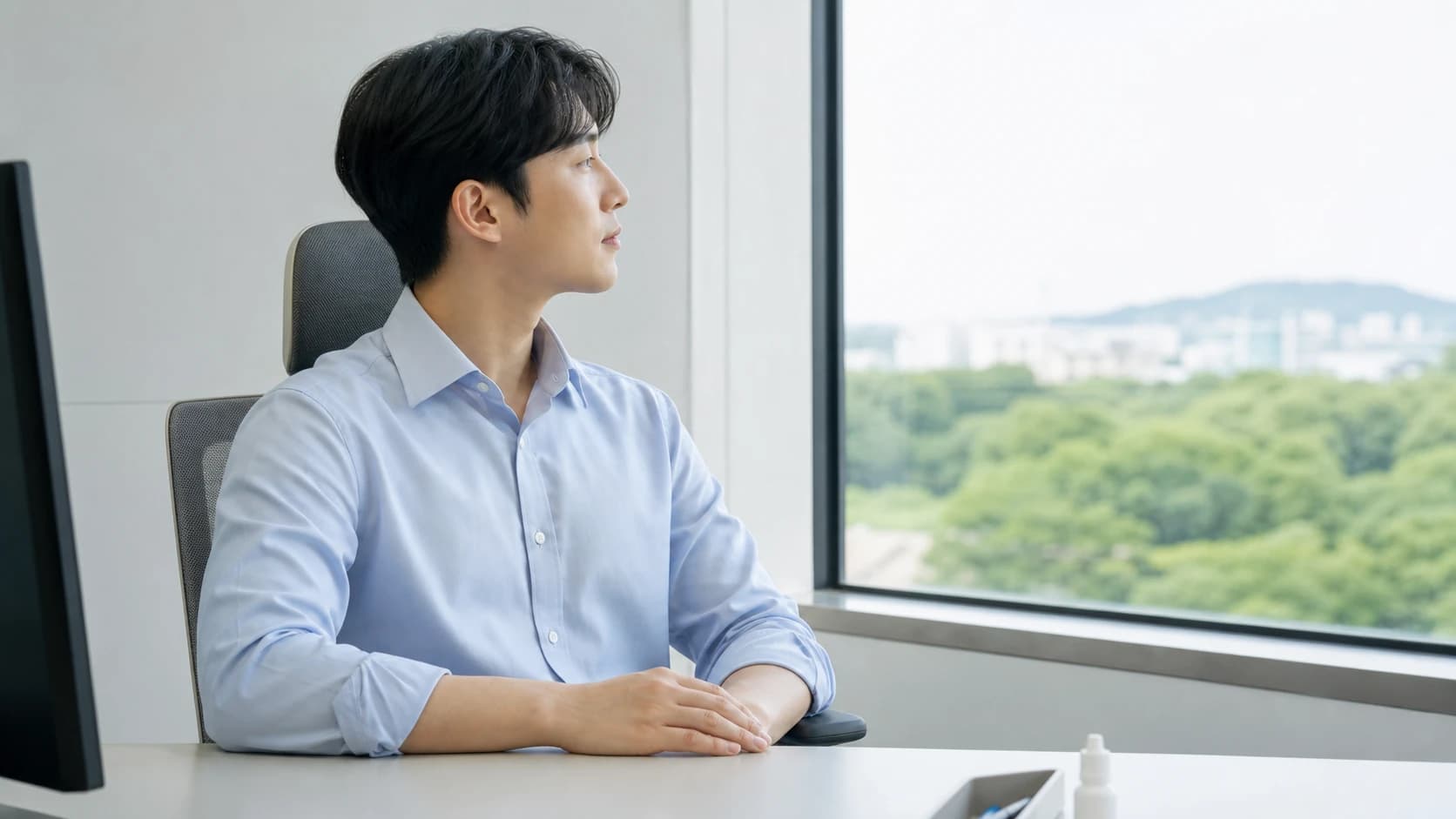 사무실에서 모니터에서 시선을 떼고 창밖을 잠시 보는 사람과 책상 위 라벨 없는 작은 점안 용기