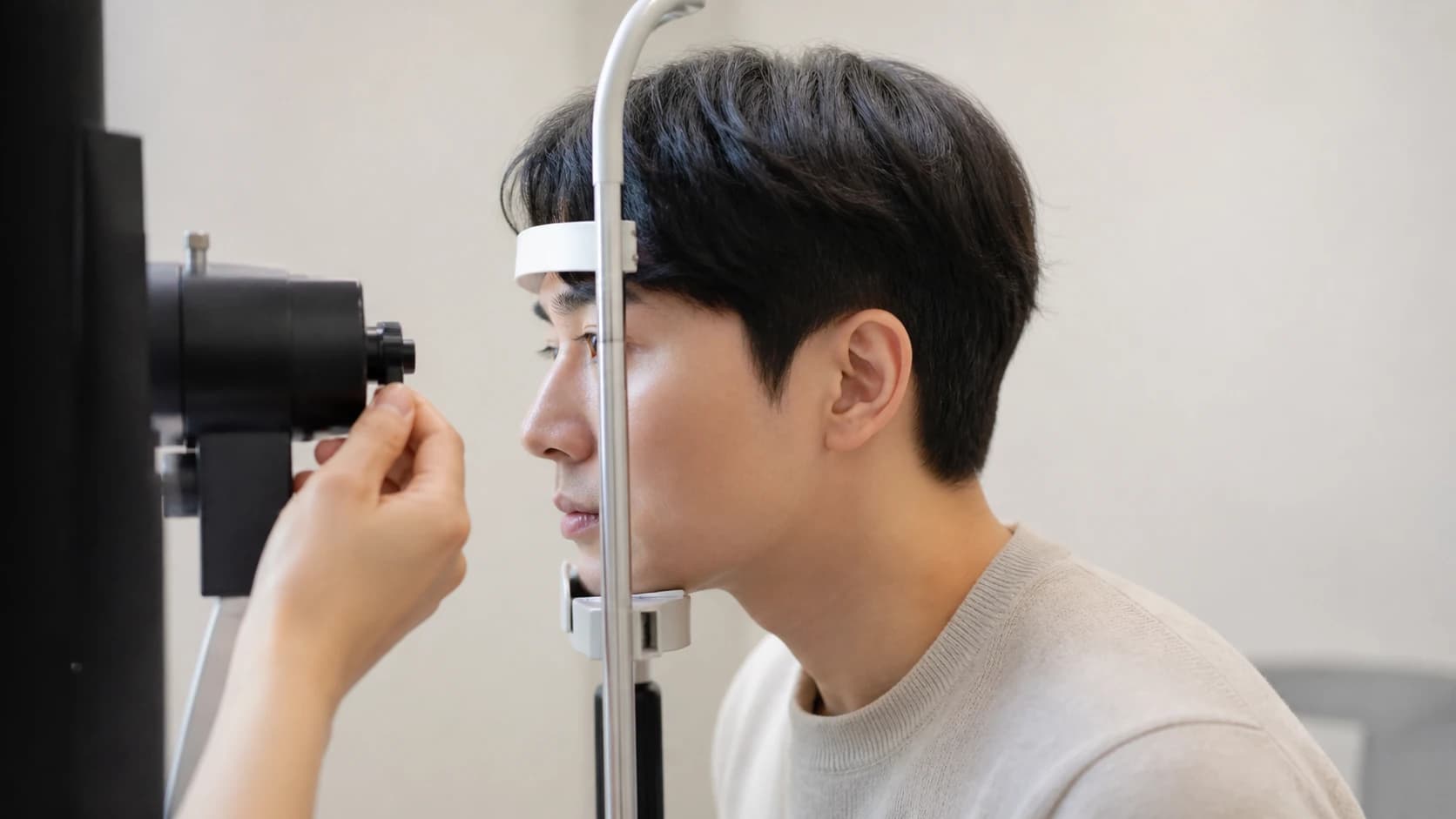 검사 장비 앞에 앉아 이마와 턱을 받침에 대고 있는 사람과 조작부에 걸친 검사자의 손