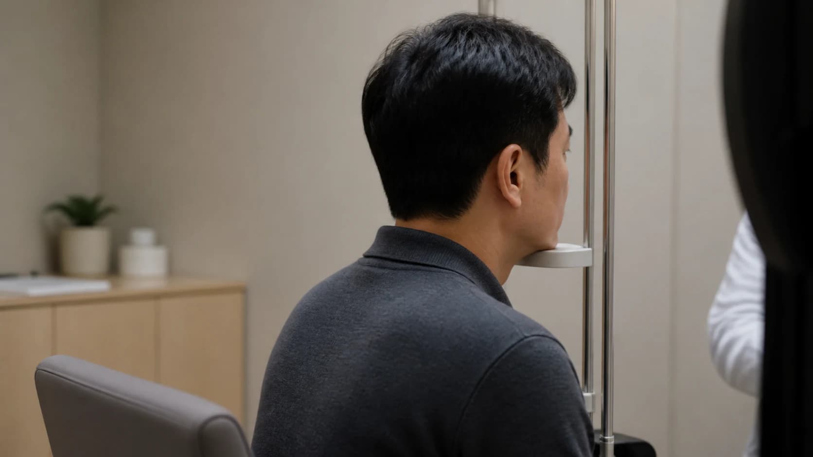 검사실에서 턱받침에 턱을 대고 정면을 응시하는 사람의 측후면 중거리, 장비는 화면 가장자리에 실루엣으로만 걸친 장면